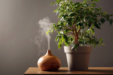 Photo of Air humidifier near houseplant on wooden table against grey wall. Space for text