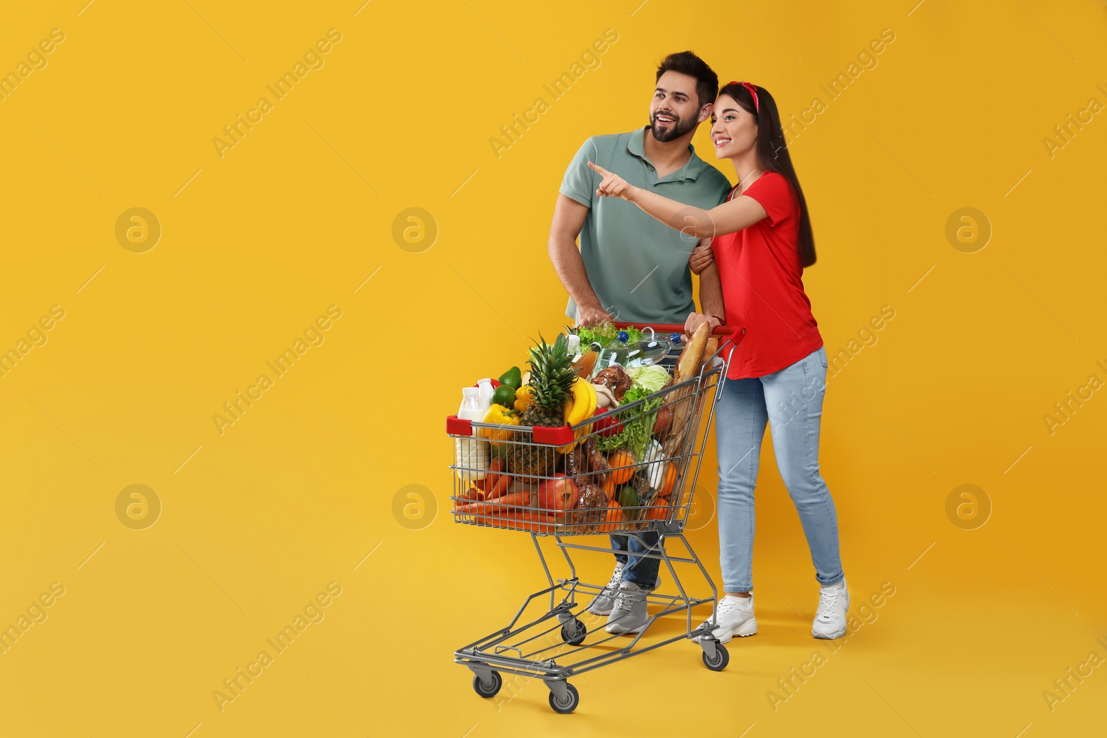 Photo of Young couple with shopping cart full of groceries on yellow background