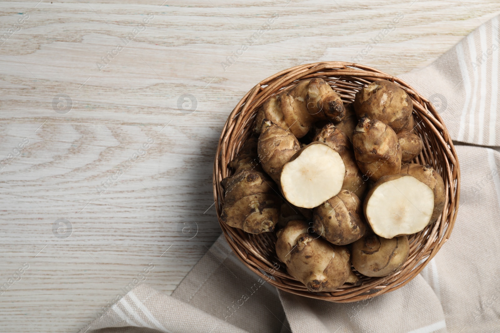 Photo of Wicker basket with many Jerusalem artichokes on white wooden table, top view. Space for text