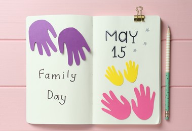 Photo of Notebook with text Family Day May 15 and paper cutouts on pink wooden table, flat lay