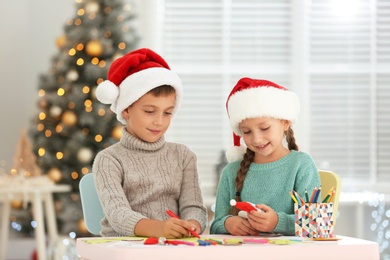 Photo of Little children in Santa hats making craftworks at table indoors. Christmas season