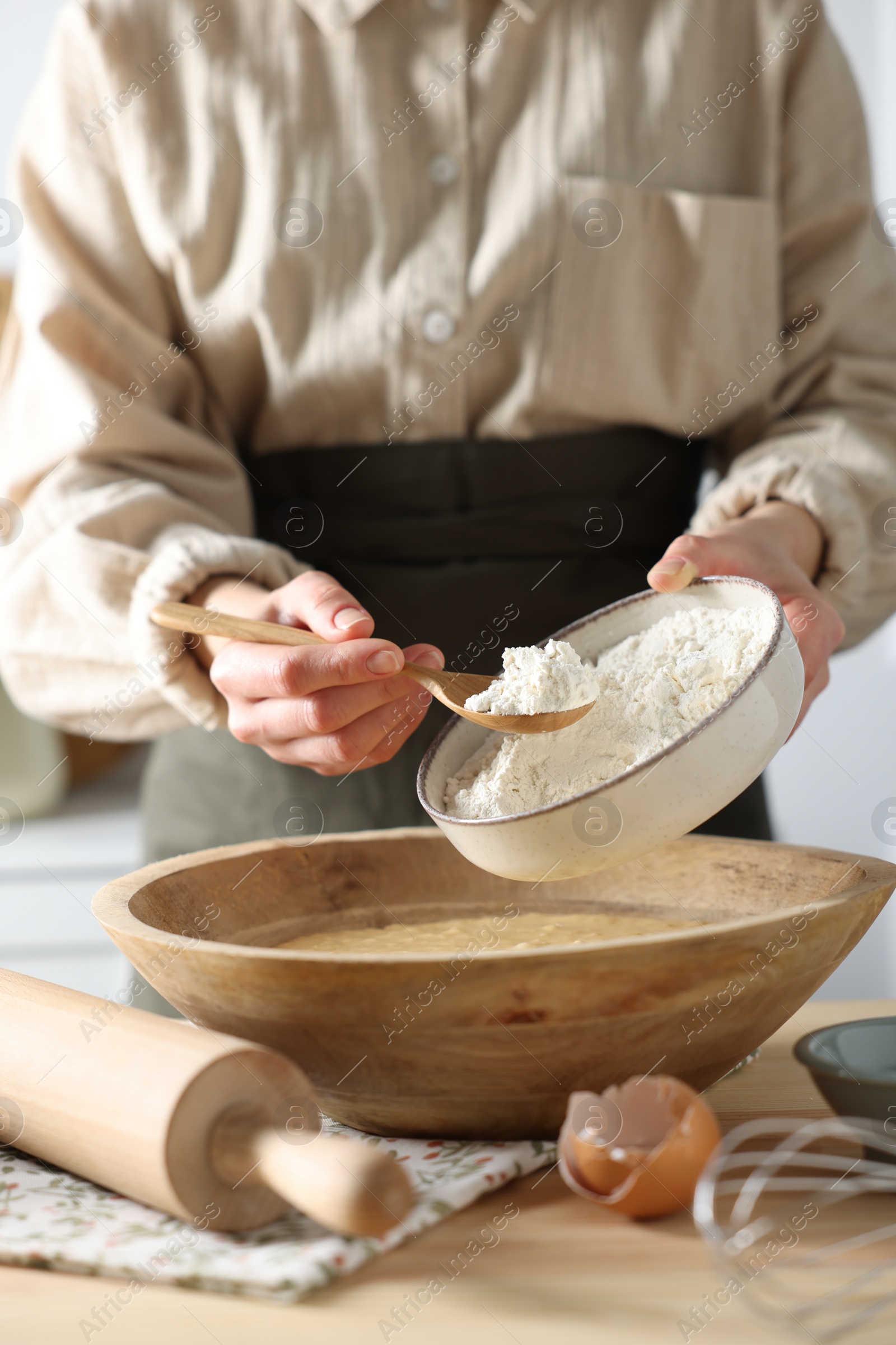 Photo of Making dough. Woman adding flour into bowl at wooden table in kitchen, closeup