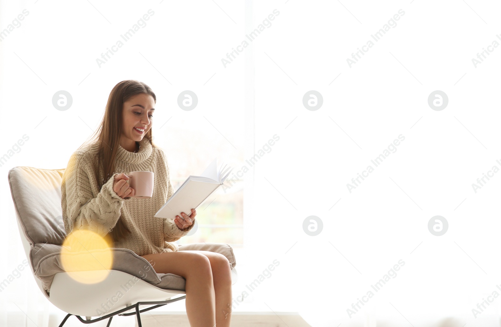 Photo of Young woman with cup of coffee reading book near window at home, space for text