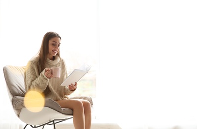 Photo of Young woman with cup of coffee reading book near window at home, space for text