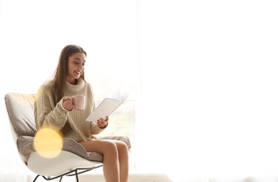 Young woman with cup of coffee reading book near window at home, space for text