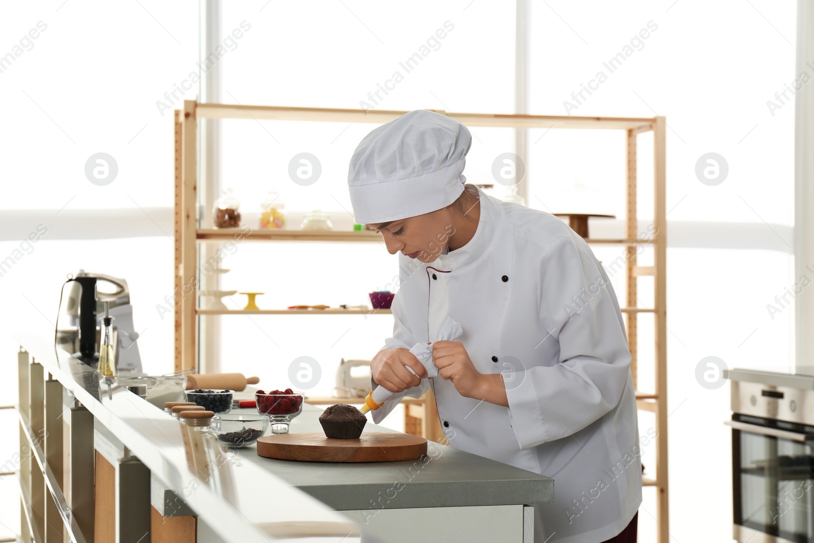 Photo of Young female pastry chef decorating cupcake with cream at table in kitchen