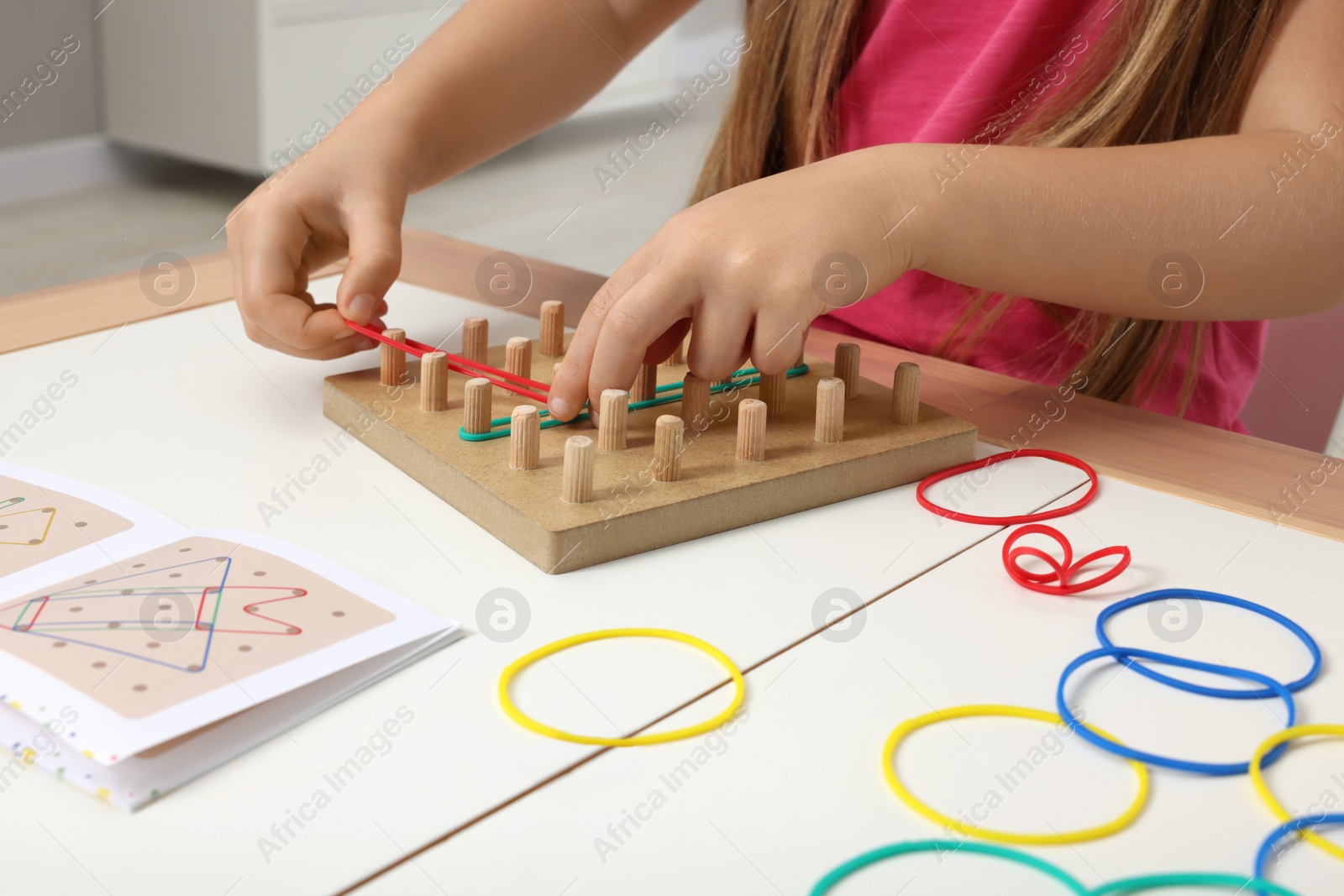 Photo of Motor skills development. Girl playing with geoboard and rubber bands at white table indoors, closeup