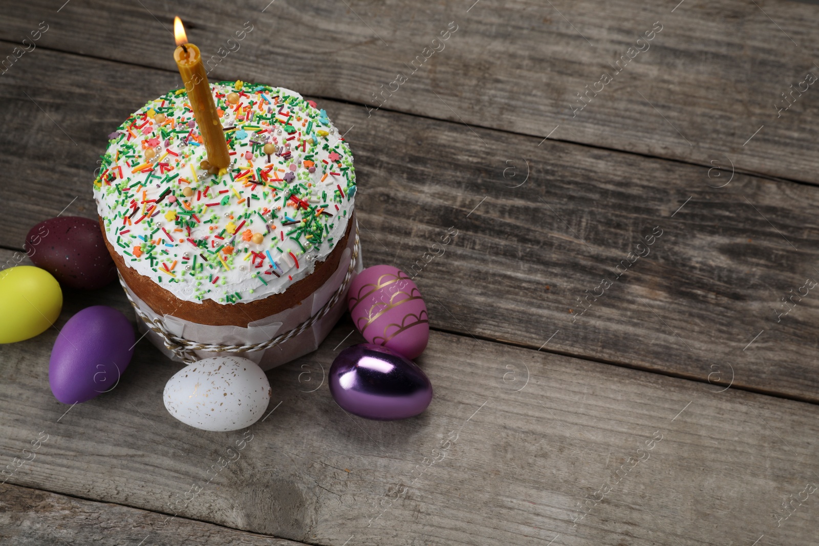 Photo of Traditional Easter cake with sprinkles, burning candle and painted eggs on wooden table, above view. Space for text