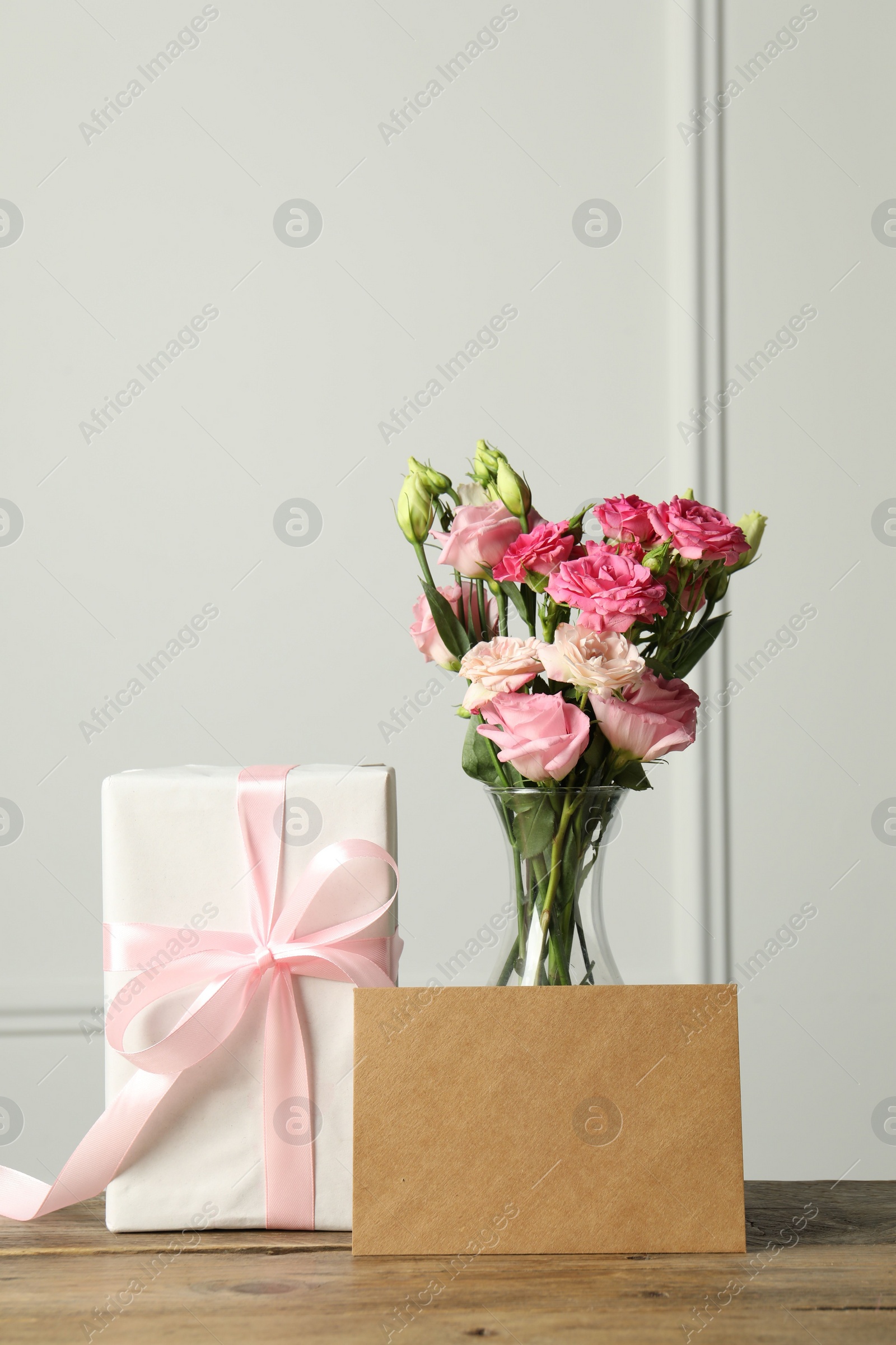 Photo of Happy Mother's Day. Gift box, blank card and bouquet of beautiful flowers in vase on wooden table near white wall