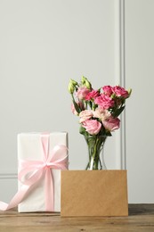 Happy Mother's Day. Gift box, blank card and bouquet of beautiful flowers in vase on wooden table near white wall