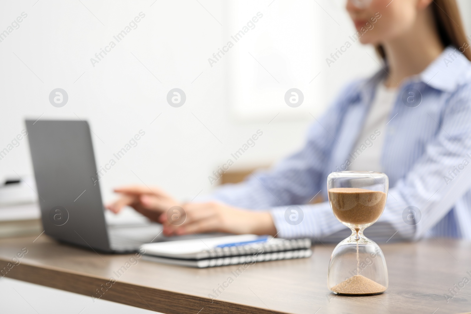 Photo of Hourglass with flowing sand on desk. Woman using laptop indoors, selective focus