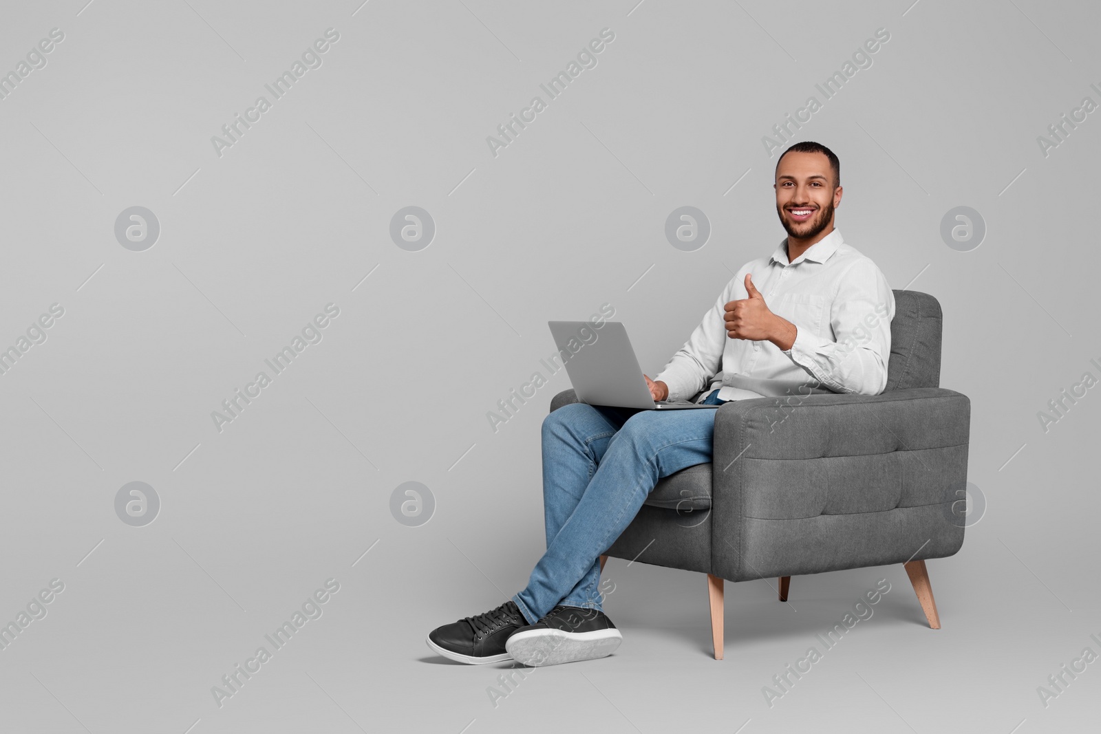 Photo of Smiling young man with laptop sitting in armchair on grey background. Space for text