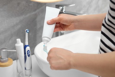 Woman squeezing toothpaste from tube onto toothbrush near sink in bathroom, closeup