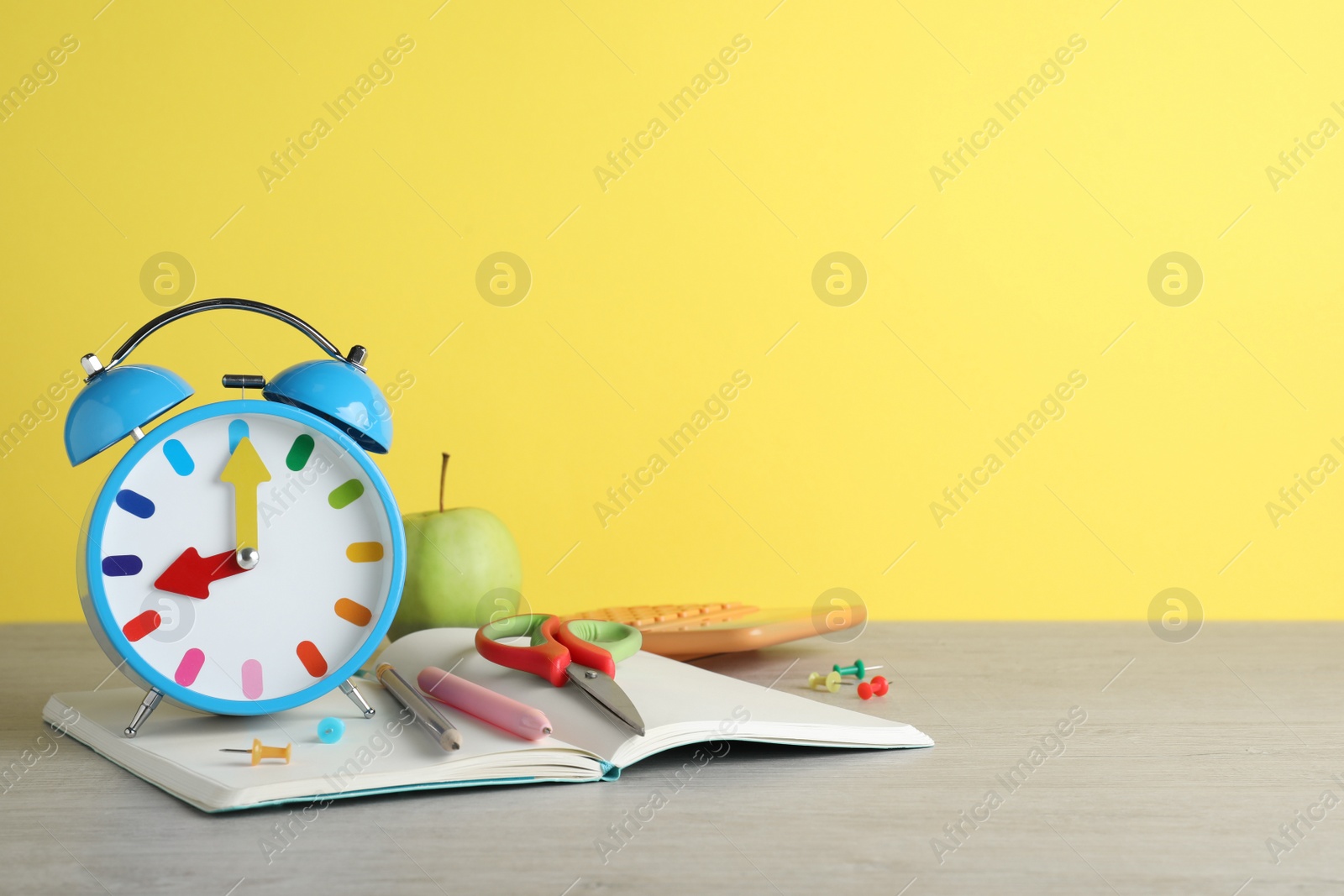 Photo of Light blue alarm clock and different stationery on white wooden table against yellow background, space for text. School time