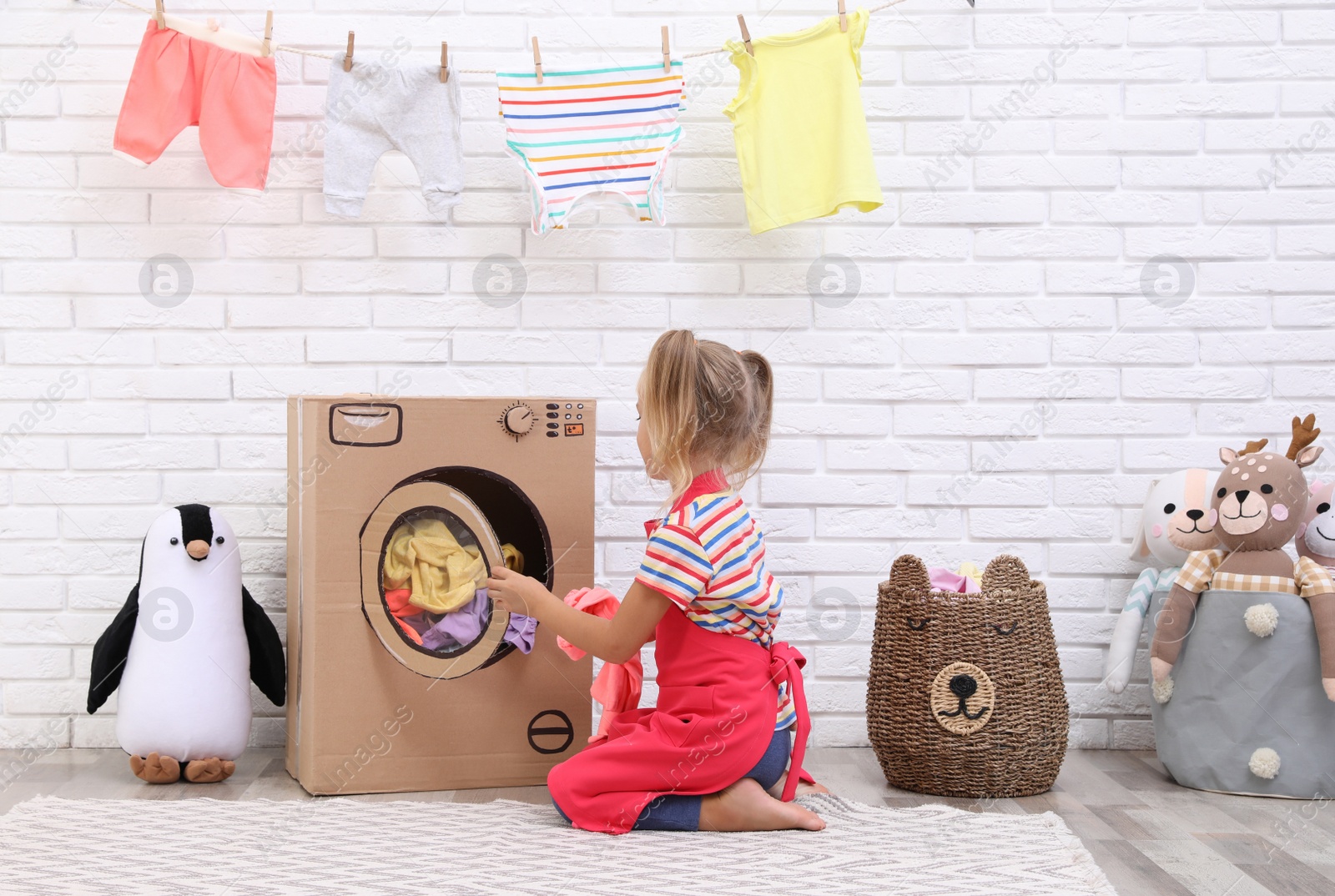 Photo of Little girl putting laundry into toy cardboard washing machine indoors