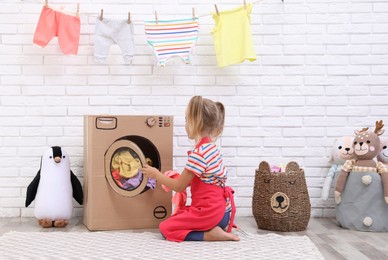 Little girl putting laundry into toy cardboard washing machine indoors