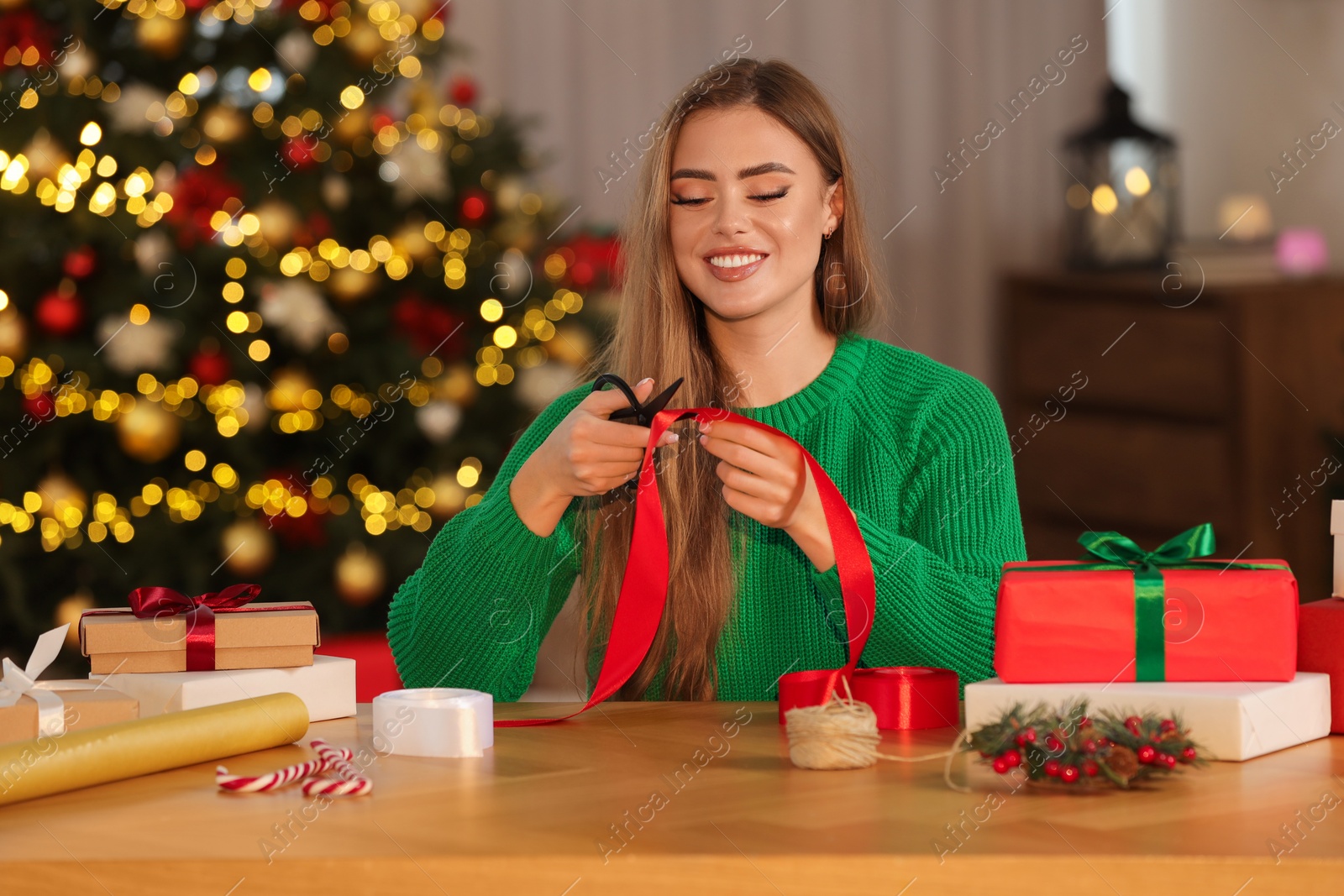 Photo of Beautiful young woman cutting red ribbon at table in room. Decorating Christmas gift