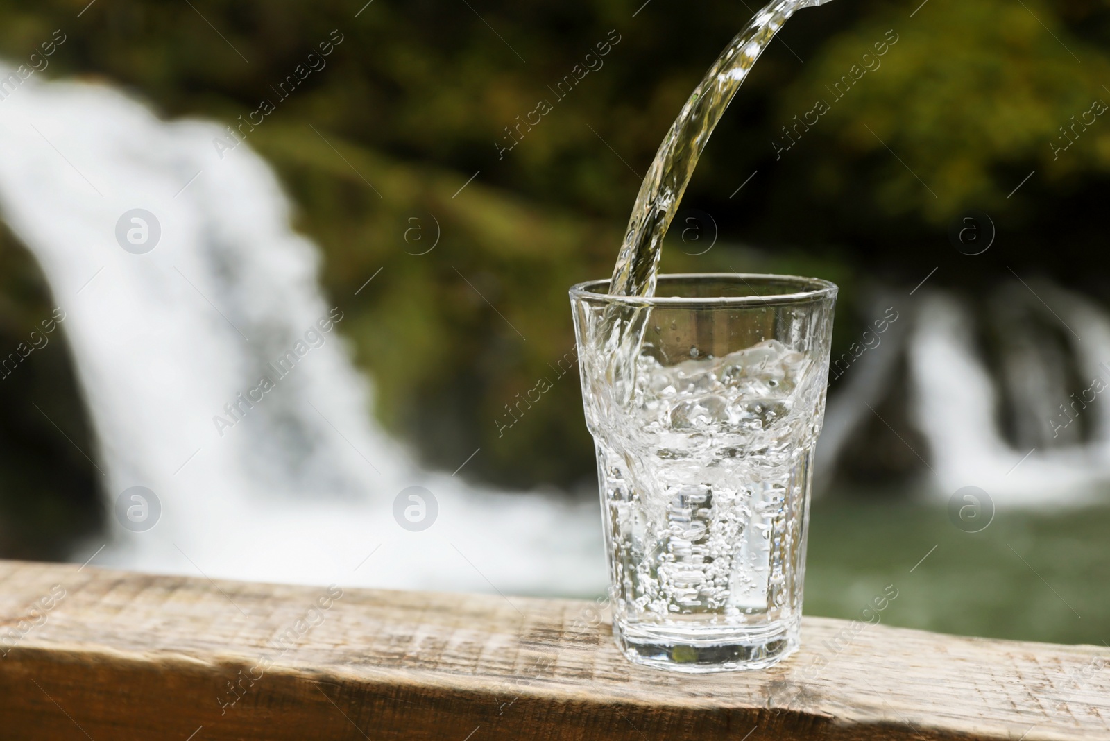 Photo of Fresh water pouring into glass on wooden surface near waterfall. Space for text