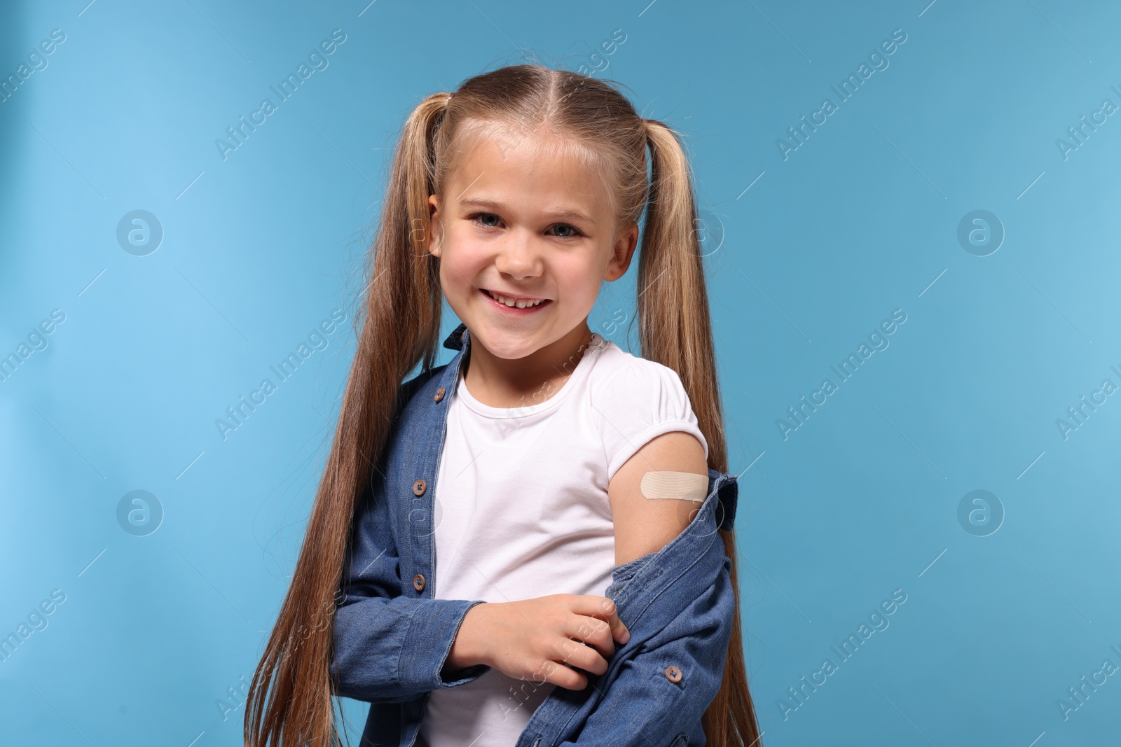 Photo of Happy girl with sticking plaster on arm after vaccination against light blue background