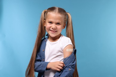 Happy girl with sticking plaster on arm after vaccination against light blue background
