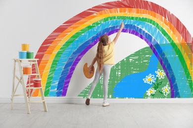 Image of Cute child drawing rainbow on white wall indoors