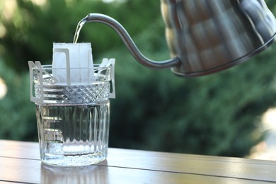 Pouring hot water into glass with drip coffee bag from kettle on wooden table, closeup