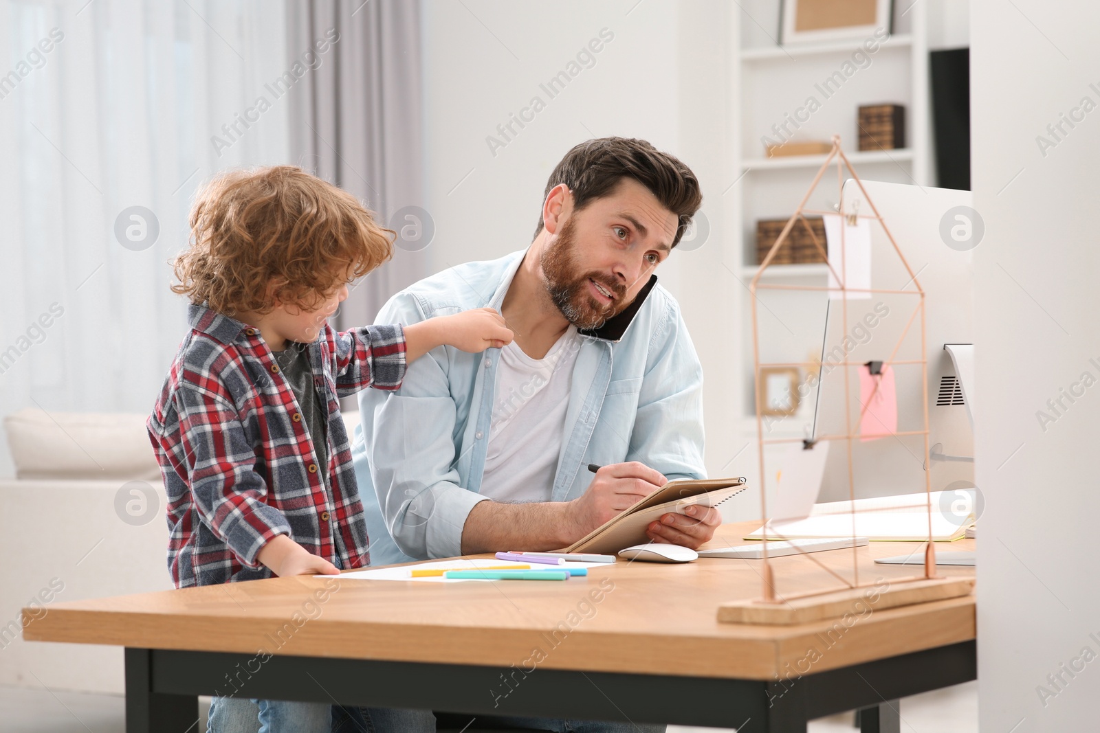 Photo of Little boy bothering his father at home. Man working remotely at desk