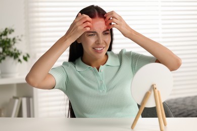 Photo of Suffering from allergy. Young woman looking in mirror and scratching her forehead at white table indoors