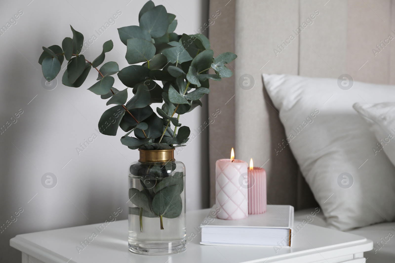 Photo of Vase with beautiful eucalyptus branches, book and candles on nightstand in bedroom