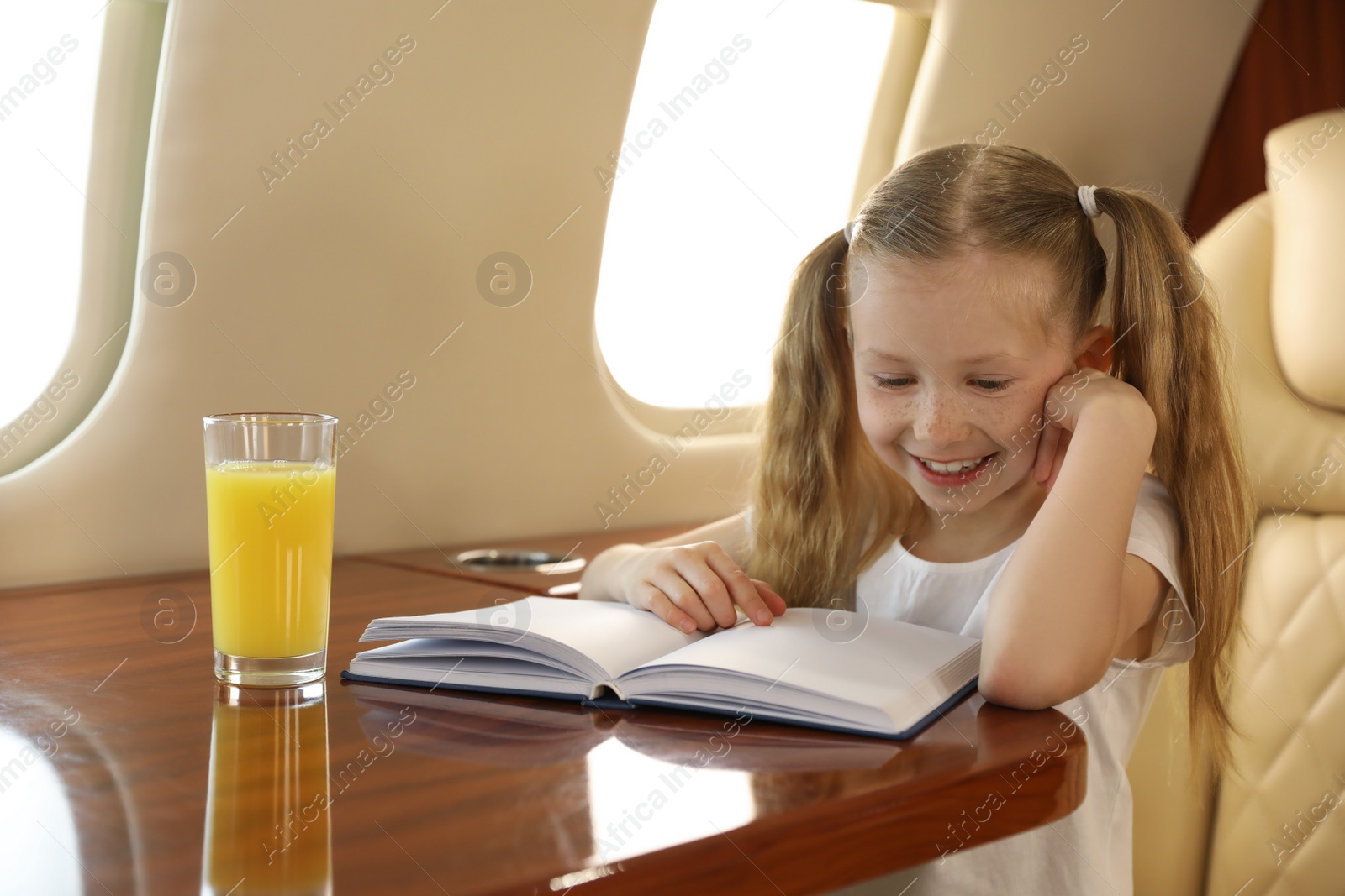 Photo of Cute little girl reading book at table in airplane during flight