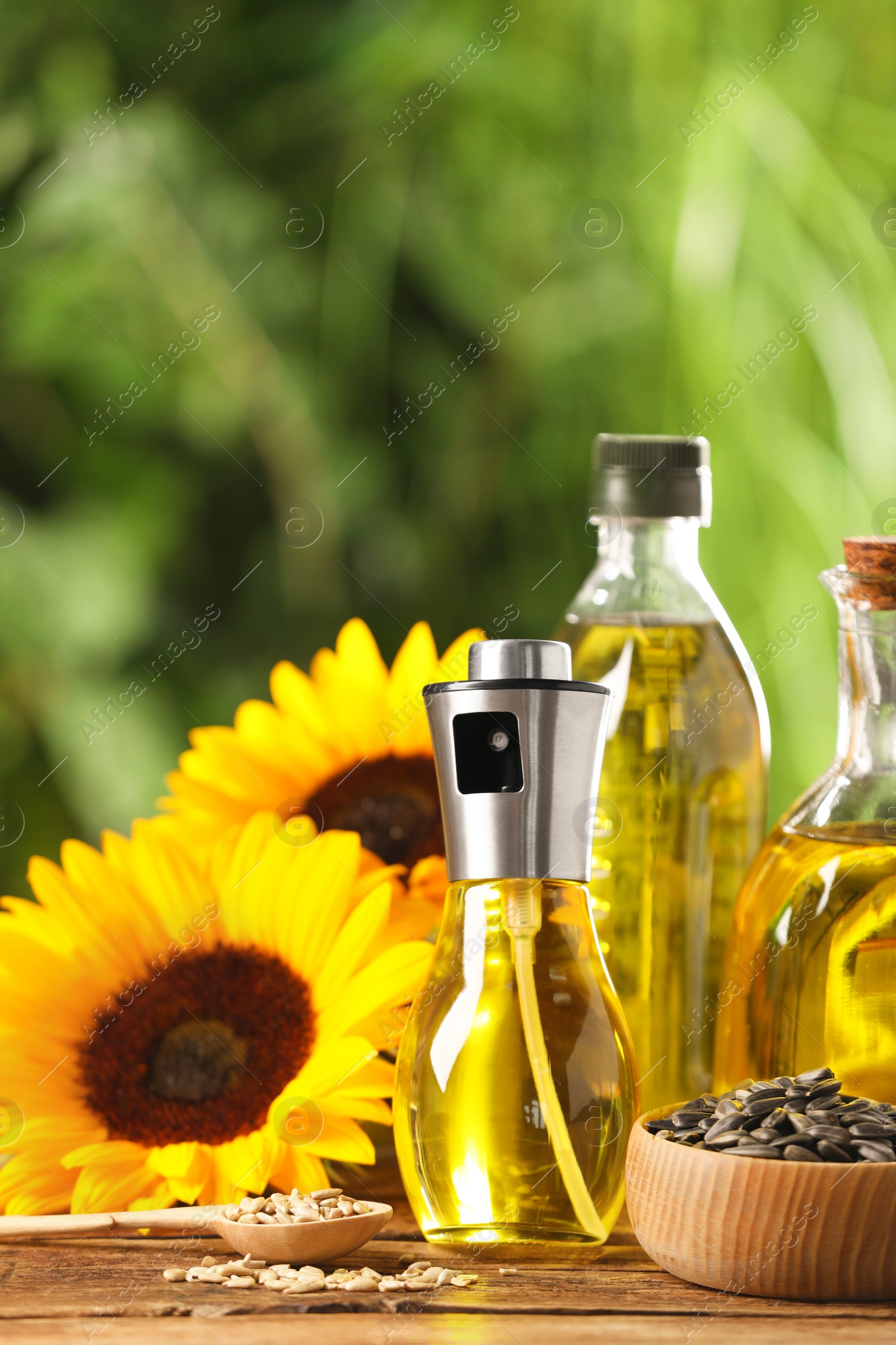 Photo of Many different bottles with cooking oil, sunflower seeds and flowers on wooden table against blurred background