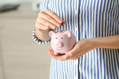 Woman putting coin into piggy bank indoors, closeup. Money savings