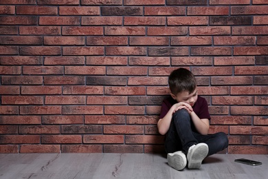 Photo of Sad little boy with mobile phone sitting on floor near brick wall. Space for text