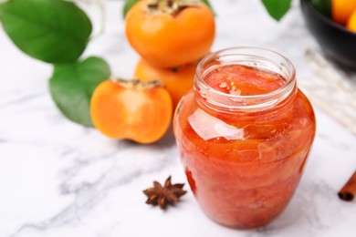 Jar of tasty persimmon jam and ingredients on white marble table. Space for text