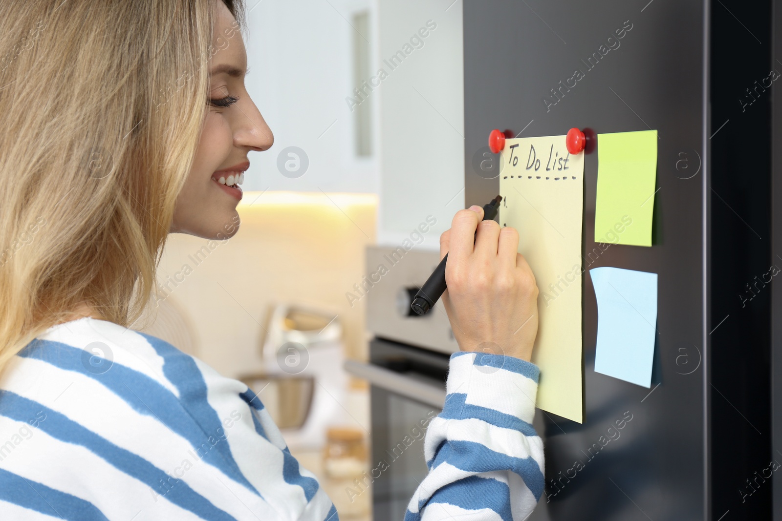 Photo of Young woman writing To do list on refrigerator door in kitchen