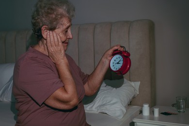 Photo of Elderly woman suffering from insomnia looking at time on alarm clock in bedroom
