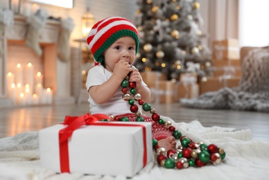Cute little baby with elf hat and decor near Christmas gift on floor at home
