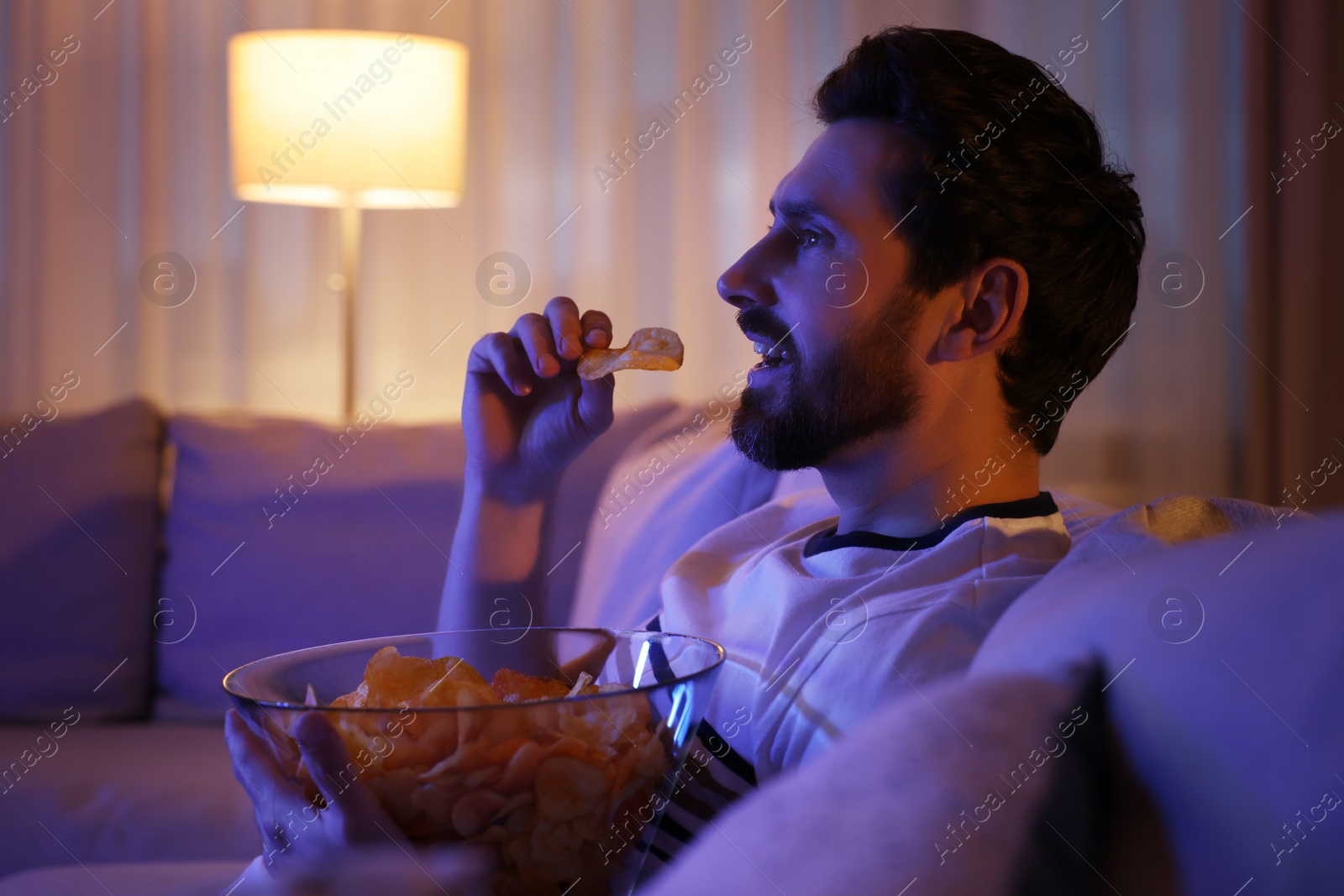 Photo of Man eating chips while watching TV on sofa at night. Bad habit