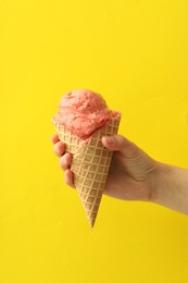 Woman holding waffle cone with delicious pink ice cream on yellow background, closeup