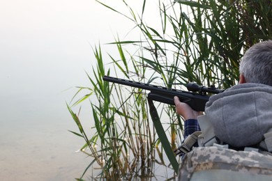 Photo of Man aiming with hunting rifle near lake outdoors, closeup. Space for text