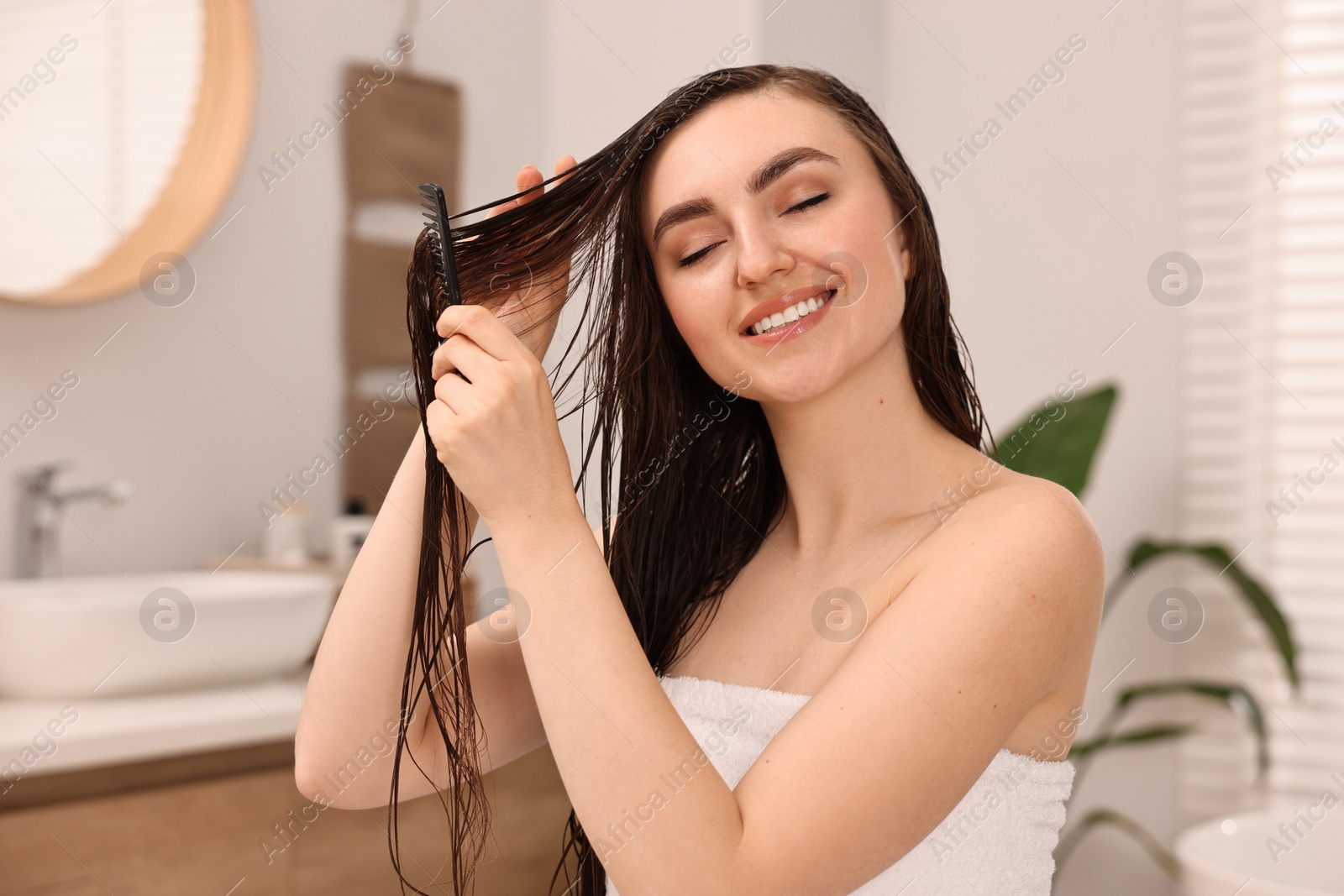 Photo of Young woman brushing hair after applying mask in bathroom