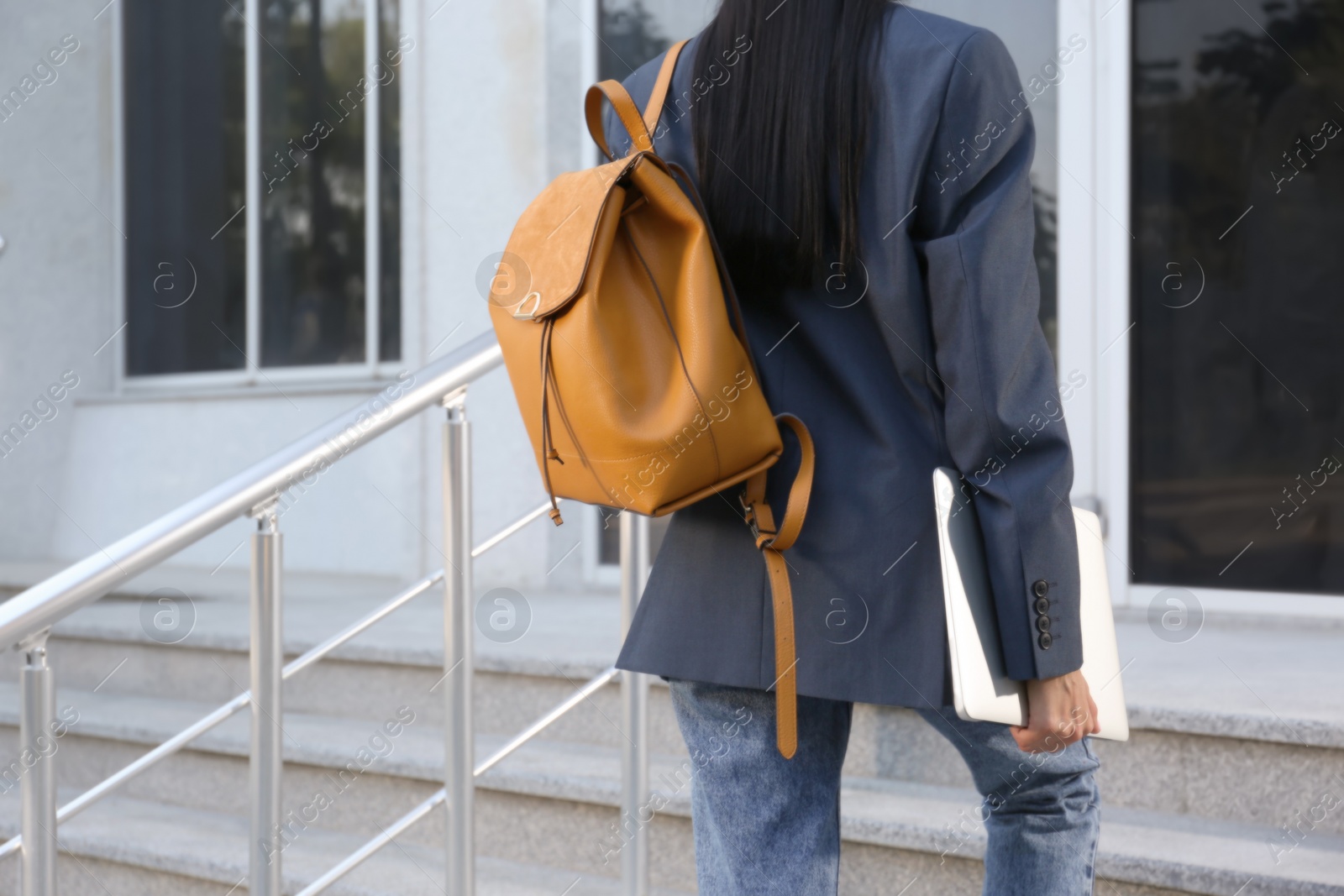 Photo of Young woman with stylish backpack and laptop going upstairs outdoors, closeup. Space for text