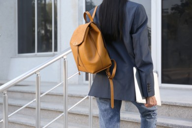Photo of Young woman with stylish backpack and laptop going upstairs outdoors, closeup. Space for text