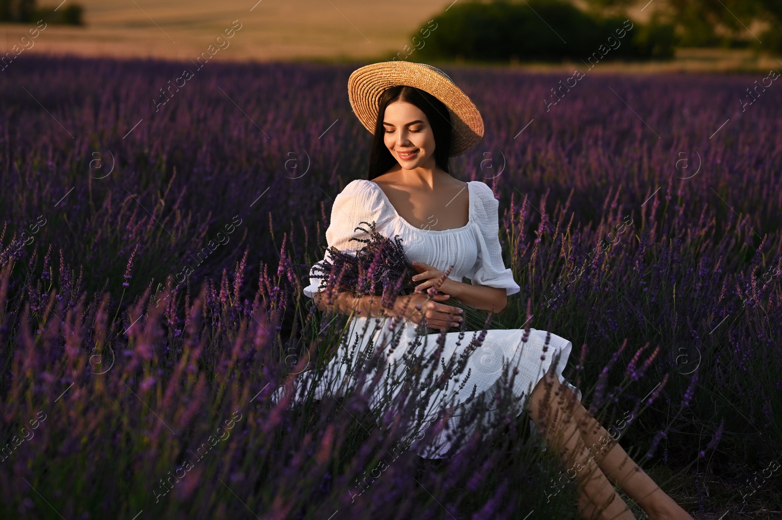 Photo of Beautiful young woman with bouquet sitting in lavender field at sunset