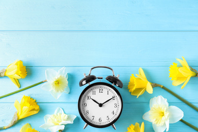 Photo of Black alarm clock with spring flowers and space for text on light blue wooden background, flat lay. Time change