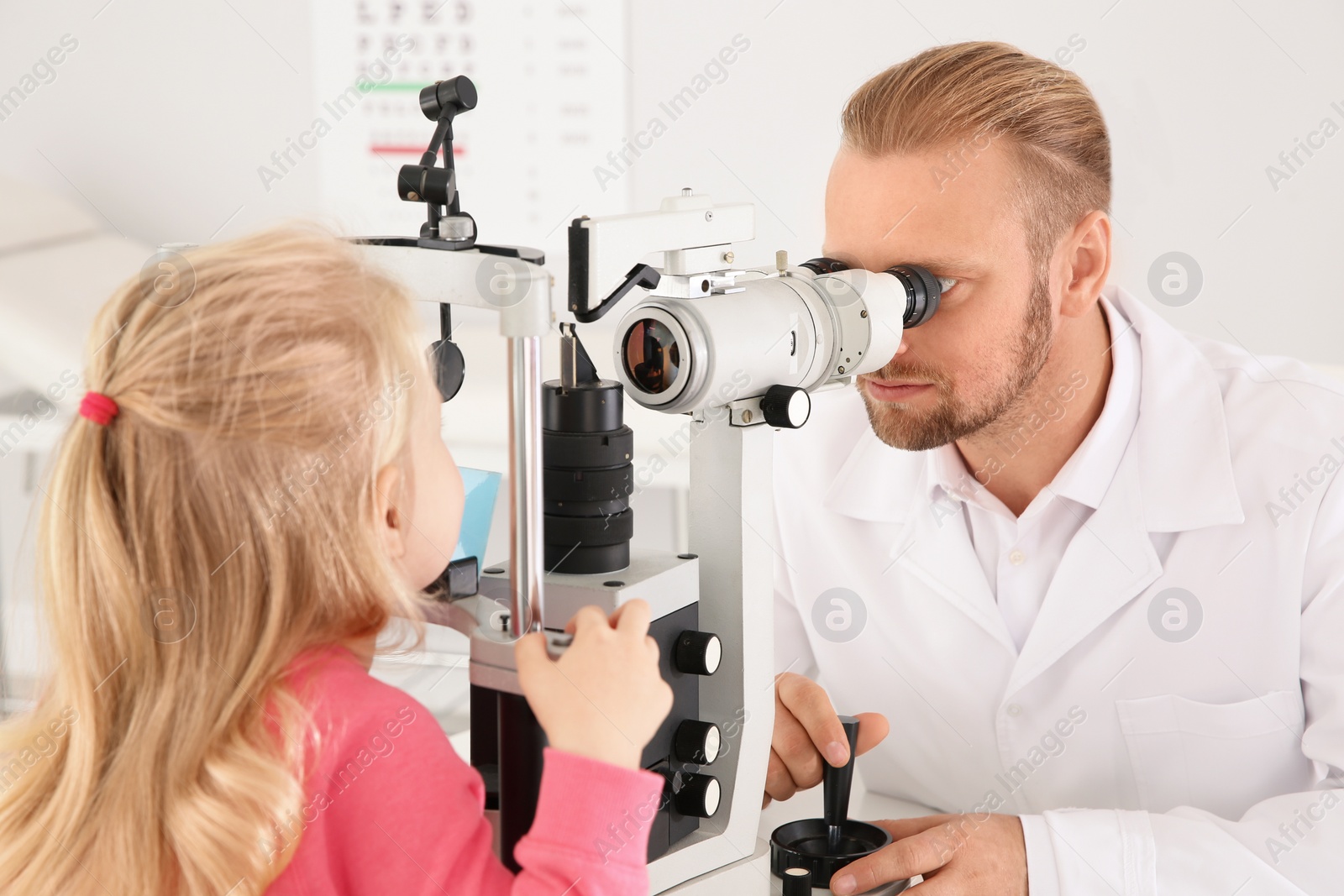 Photo of Children's doctor examining little girl with ophthalmic equipment in clinic