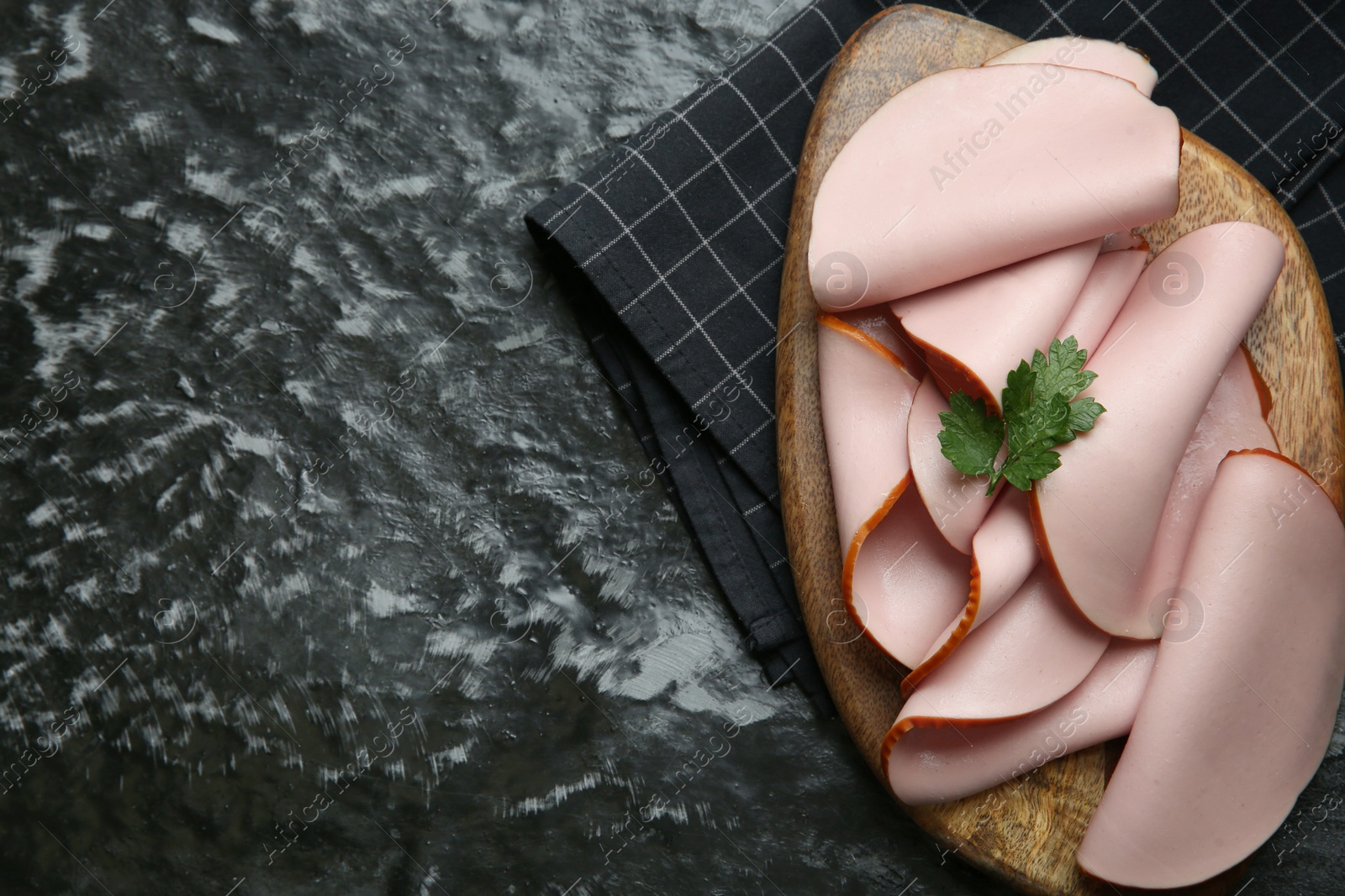 Photo of Board with slices of tasty boiled sausage and parsley on dark textured table, top view. Space for text