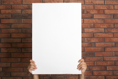Photo of Man holding white blank poster near red brick wall, closeup. Mockup for design