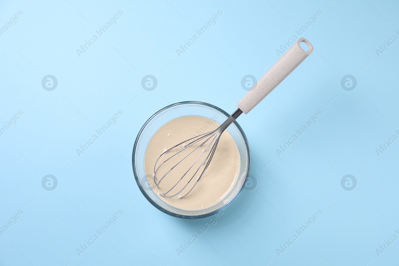 Photo of Dough and whisk in glass bowl on light blue background, top view