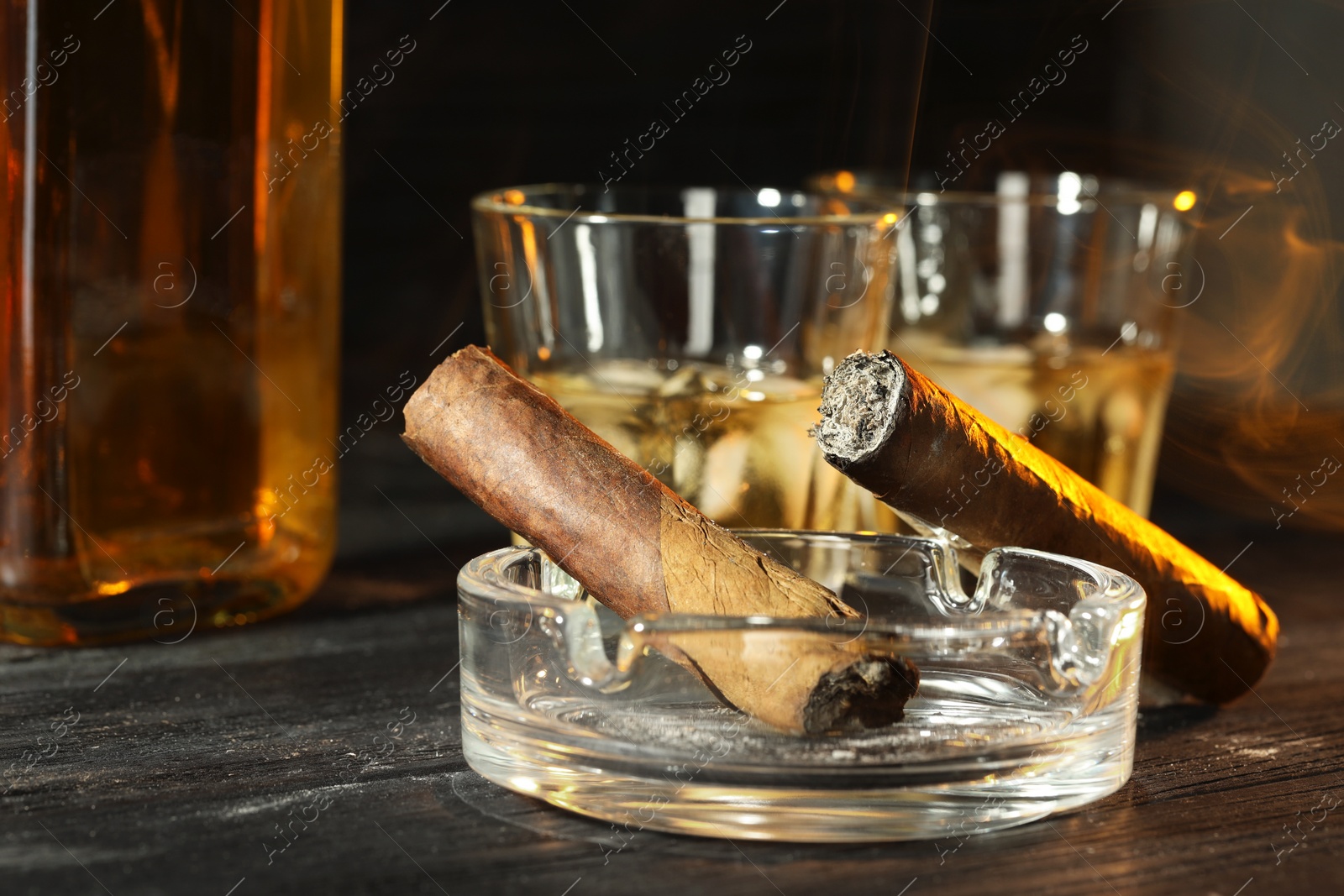 Photo of Cigars, ashtray and whiskey with ice cubes on black wooden table, closeup